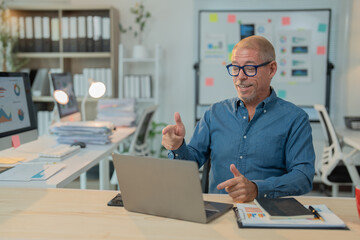 Cheerful mature entrepreneur gesturing and making video conference with colleagues on laptop, discussing project strategy during online meeting in modern workplace