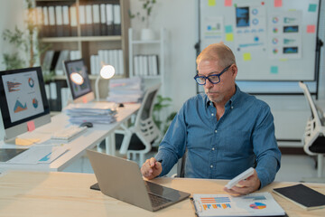 Focused mature manager analyzing marketing data using laptop computer and holding smartphone while sitting at workplace in modern office with documents and graphs on table