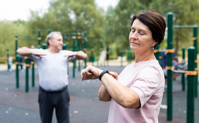 Mature woman in sportswear looks at fitness watch or at pedometer while exercising in city park