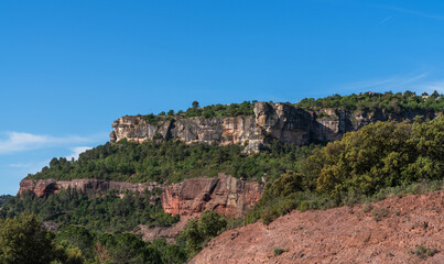 Fototapeta premium A serene landscape unfolds with a rugged, reddish-brown earth foreground leading to lush greenery, crowned by towering cliffs under a clear blue sky.