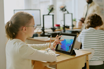 Girl engaging with interactive tablet in modern classroom setting, wearing glasses while seated at a desk, with classmates in background in a collaborative learning environment
