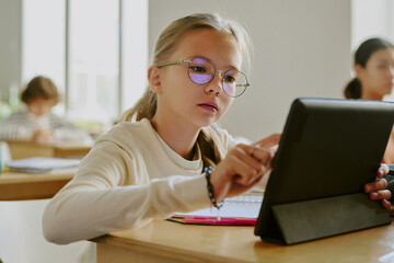 Young girl wearing glasses sitting at wooden desk using tablet in classroom surrounded by fellow students in bright learning environment