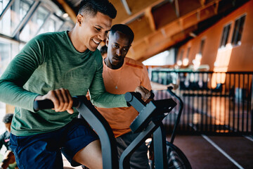 Smiling trainer offering encouragement to a fit young man using a stationary bicycle during a workout session in a gym