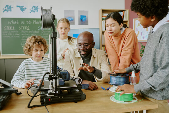 Diverse group of students gathered around a teacher explaining 3D modeling using a 3D printer in classroom setting with educational posters on walls