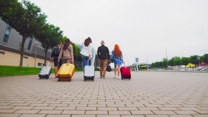 Group of travelers walking with luggage at modern airport. A group of five travelers walks down a paved path towards a modern airport building, each carrying a suitcase.