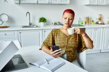 A bald woman with vibrant red hair sips coffee while gazing at her phone in a cozy kitchen.