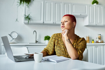 A vibrant woman thoughtfully engages with her laptop while sipping coffee in a modern kitchen.