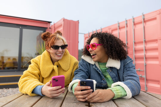 Female friends messing about wearing fun sunglasses on a beach in the winter holding their phones