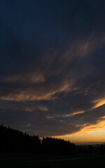 An autumn landscape at sunset, featuring a dramatic sky filled with vibrant colors, dominated by beautifully shaped clouds that are illuminated by the warm hues of the setting sun.