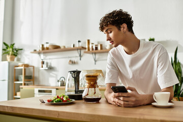 A handsome young man with curly hair sits at a table, sipping coffee while checking his phone,...