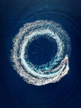 Aerial top view of a motor RIB boat forming a circle of bubbles and surf with its engine over blue ocean