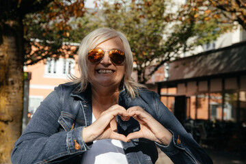 Fototapeta premium Happy woman with sunglasses making heart gesture in outdoor urban setting
