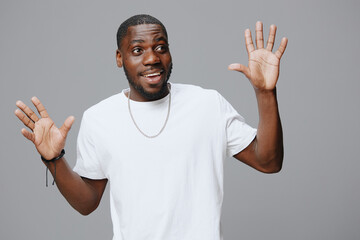 Fototapeta premium Happy young man expressing surprise with raised hands in a white t shirt, against a neutral grey background, showcasing joy and enthusiasm, ideal for diverse lifestyle concepts