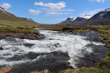 panoramic view of an icelandic landscape with a wild mountain river in a rocky terrain with numerous waterfalls coming down the mountain cliff in the distance that is stil partially covered with snow