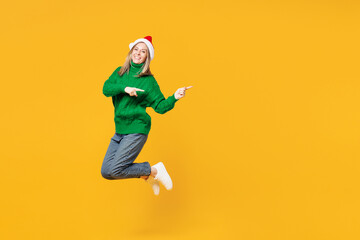 Obraz premium Full body merry young woman wears green casual sweater Santa hat posing jump high point index finger aside isolated on plain yellow background. Happy New Year, celebration Christmas holiday concept.