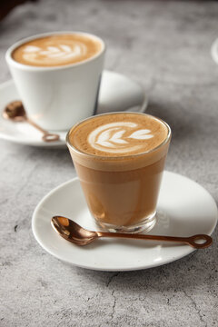 two cups of coffee with latte art on top on a white saucer with copper-coloured spoon