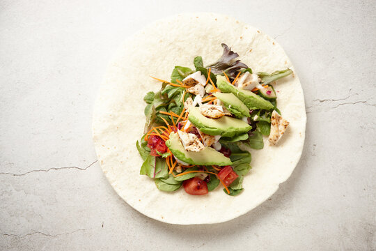 Tortilla wrap topped with spinach, avocado, tomato, chicken, and carrots on a white surface