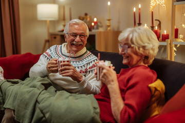 Older couple sitting comfortable on a sofa during Christmas holidays and drinking hot cocoa