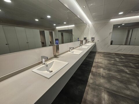 Rang&eacute;e de lavabos modernes align&eacute;s sous un grand miroir dans une salle de bain spacieuse, avec des murs clairs et un sol sombre dans un a&eacute;roport