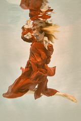 Girl in red dress with long hair swims underwater on light background