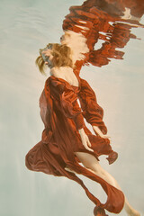 Girl in red dress with long hair swims underwater on light background
