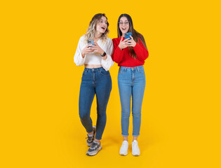 Fototapeta premium Shocked surprised happy two beautiful full body size front view young caucasian women using mobile phone looking touch screen isolated over yellow studio background. Wear casual jean and shirt.