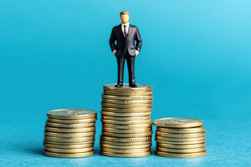 Miniature figure in a suit standing on stacks of coins, blue background.