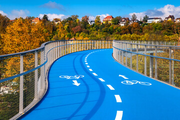 New built bicycle bridge in Tübingen (Germany) with curved, blue-colored roadway on a sunny autumn day. 2 cycle lanes with white lines and pictograms lead with incline in curves over a railroad.