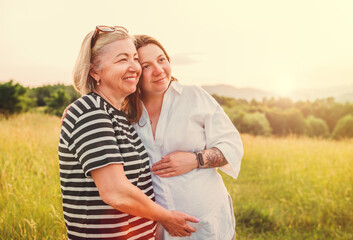 Obraz premium Portrait of a sincerely smiling young pregnant woman dressed in light summer clothes embracing with mother in evening sunset hours. Woman's health, human in the nature concept image.