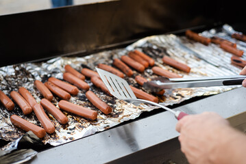 Caucasian man hand holding spatula and tongs flipping, cooking sausages on grilling tin foil sheet at outdoor BBQ party in Dallas, Texas, gas grill roasting on indirect heat cookout concept