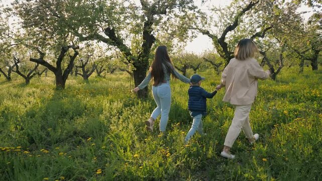 A family holding hands walks through a green apple orchard in spring, surrounded by blooming trees and vibrant grass. Sunlight filters through the branches