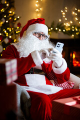 Jolly Santa Claus with eyeglasses scrolls his smartphone, reads heartfelt letters by a cozy fireplace in a cozy festive room with Christmas tree, decorated by twinkling lights. Coming holiday vibes