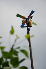 A colorful wooden windmill on a pole, producing a tin-like sound when spinning. Decorated with two cartoon figures whose arms move with each breeze-driven rotation.