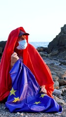 Migrant in Europe with the sea in the background. The European Community flag held by an illegal immigrant wearing a red first aid blanket, a protective mask and a hat