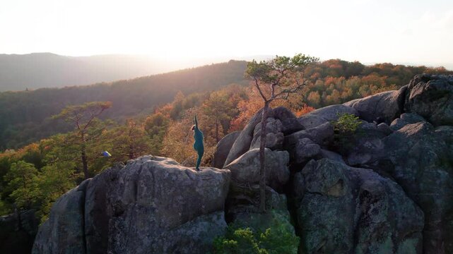 Woman practicing yoga and meditation on the top of huge boulder in the evening. Beautiful sunset, autumn forests, rocks and hills on background. Aerial drone view