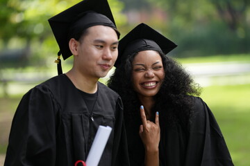 Two graduates in caps and gowns joyfully celebrate, each holding a diploma with smiles and raised hands. Their excitement shines in an outdoor park with greenery, marking a proud moment of achievement