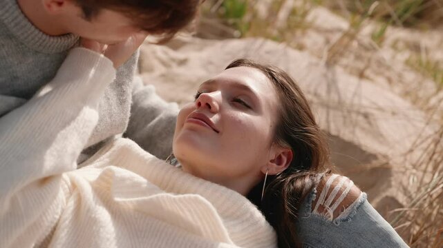 A young couple in love sharing a tender moment, with the woman lying on a man's lap, both dressed in cozy sweaters amidst a serene beach setting.