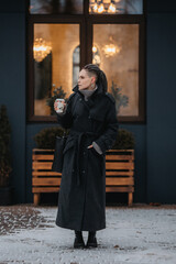 A girl with dreadlocks and piercings in a coat is drinking coffee while warming herself on the...