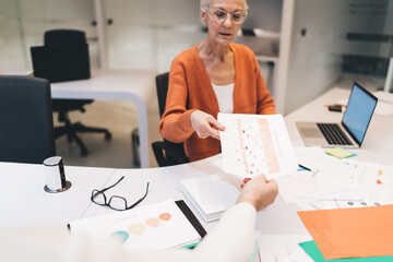 Senior businesswoman handing printed chart to her colleague during discussion. desk with papers, documents, and a laptop, representing a collaborative work environment focused on data analysis
