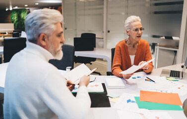 Senior woman in orange cardigan discusses paperwork with an older male colleague in a modern office...