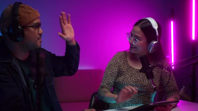 Two young creator radio hosts in headphone reviewing script to record podcast and cheering up teamwork at home studio broadcast room, cheerful cohost ready and giving high five to stream live online.
