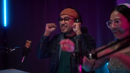 Smiling two young creator radio podcast in headphone being live streaming and speaking using microphones at studio broadcast, cheerful vloggers broadcast speaking to audiences and giving high five. 
