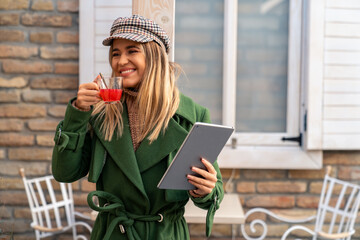 A cheerful Caucasian woman in a green coat sips a hot drink and holds a digital tablet, delighting...