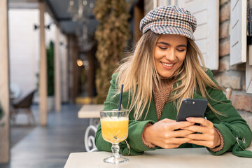 A cheerful woman relaxes with a warm beverage, browsing her smartphone at an outdoor cafe, dressed...