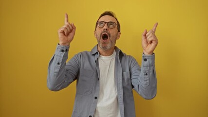 Hispanic man middle age wearing glasses standing and pointing up looking amazed over isolated yellow background, surprised expression, finger pointing, happy mood
