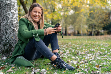 A joyful Caucasian woman in casual green outerwear sits by a tree, engaged with her smartphone,...