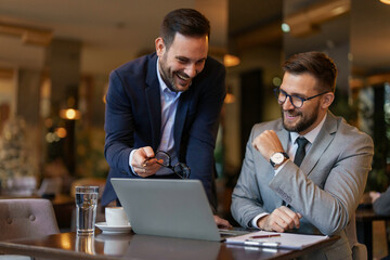 Two mid-adult Caucasian businessmen collaborate using a laptop in a trendy cafe, engrossed in work...