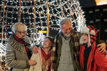 Fototapeta premium A cheerful Caucasian elderly couple enjoys a winter evening with their young granddaughter and grandson, all wrapped in warm clothes, surrounded by twinkling holiday lights.
