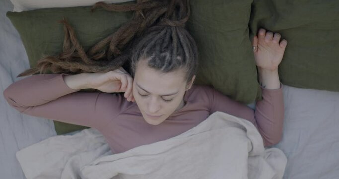 Top view of young woman with dreadlocks sleeping in bed on pillow under blanket moving in sleep enjoying rest. People and bedtime concept.