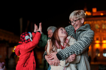 Fototapeta premium A joyful Caucasian senior woman embraces her young granddaughter, both smiling brightly, sharing a tender moment during an evening festive event, with blurred figures and lights in the background.